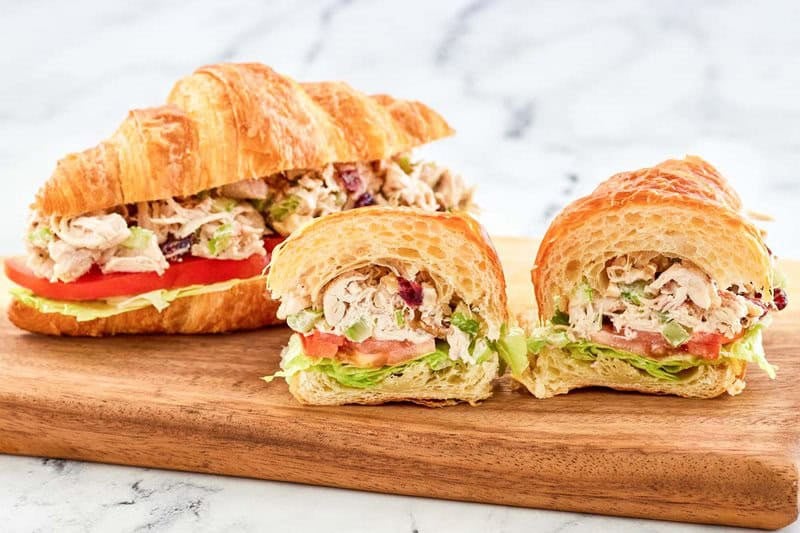 Chicken salad with cranberries and walnuts in croissant sandwiches on a wood board.