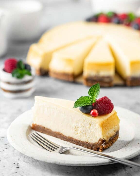 New York style cheesecake slice on a plate and the cheesecake behind it.