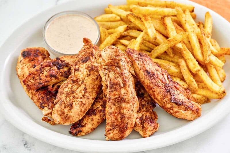 Copycat Popeyes blackened chicken tenders on a plate with fries and dipping sauce.