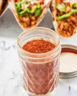 Homemade chicken taco seasoning in a jar and chicken tacos behind it.