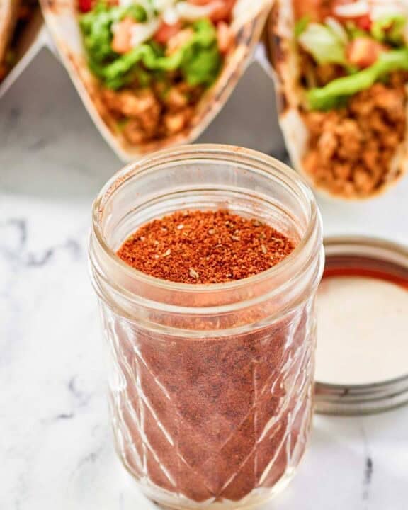 Homemade chicken taco seasoning in a jar and chicken tacos behind it.