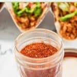 Homemade chicken taco seasoning in a jar and chicken tacos behind it.