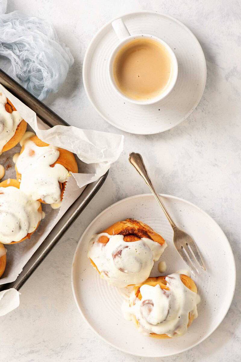 Two homemade cinnamon rolls with maple cream cheese frosting, a cup of coffee, and the rolls in a pan.