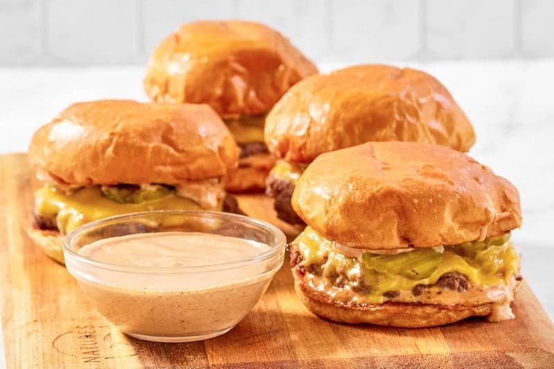 A bowl of copycat Sonic smasher sauce and smasher burgers on a wood board.