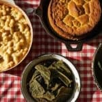 Overhead shot of a vintage Southern potluck spread on a red and white checkered tablecloth, bathed in warm, natural light. The spread features creamy mac and cheese in a rustic, slightly browned baking dish, a golden-brown cornbread in a cast iron skillet with visible texture, and vibrant green collard greens in a chipped enamel bowl showing signs of age. The scene emphasizes homey comfort and abundant hospitality, photographed with sharp focus and natural lighting.
