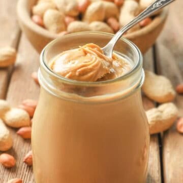 Homemade peanut butter in a glass jar and a bowl of peanuts behind it.