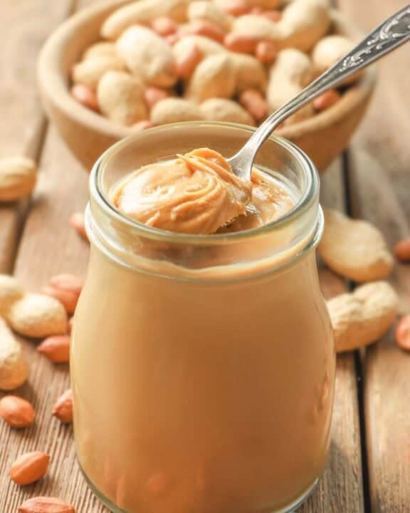 Homemade peanut butter in a glass jar and a bowl of peanuts behind it.