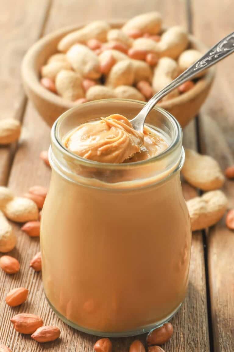 Homemade peanut butter in a glass jar and a bowl of peanuts behind it.