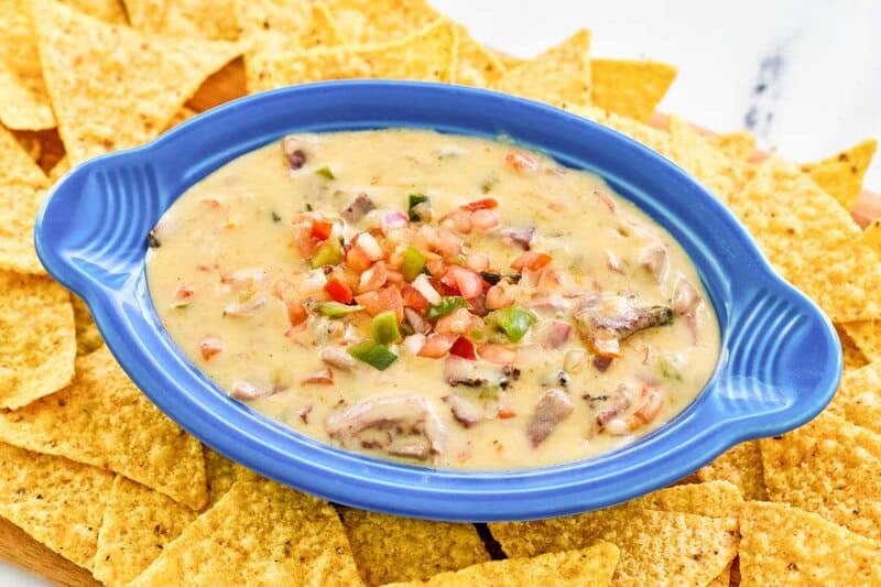 A blue oval dish filled with creamy, cheesy HEB brisket queso, featuring visible chunks of smoked brisket and fresh pico de gallo. The dish is surrounded by a generous amount of tortilla chips.