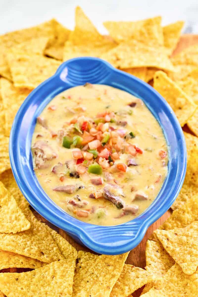 A close-up, angled shot of a blue bowl of HEB brisket queso. The image highlights the creamy texture of the cheese dip, which is loaded with chunks of smoked brisket and topped with pico de gallo, all surrounded by tortilla chips.