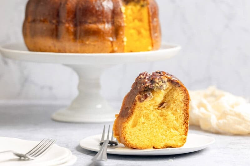 Homemade Bacardi rum cake slice on a plate and the cake on a cake stand behind it.