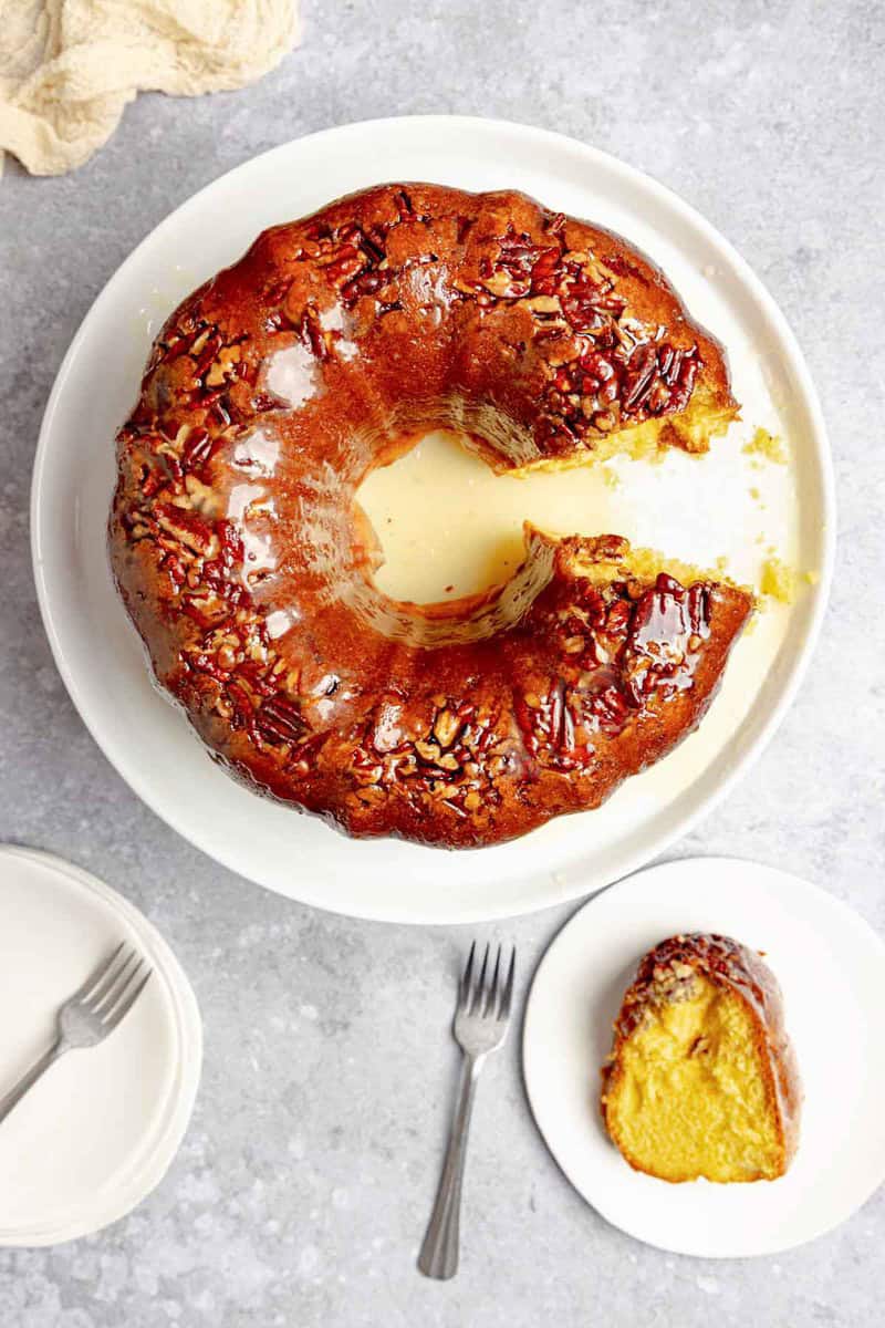 An overhead view of a glazed pecan rum cake on a platter with one slice removed. The slice sits on a separate small plate.