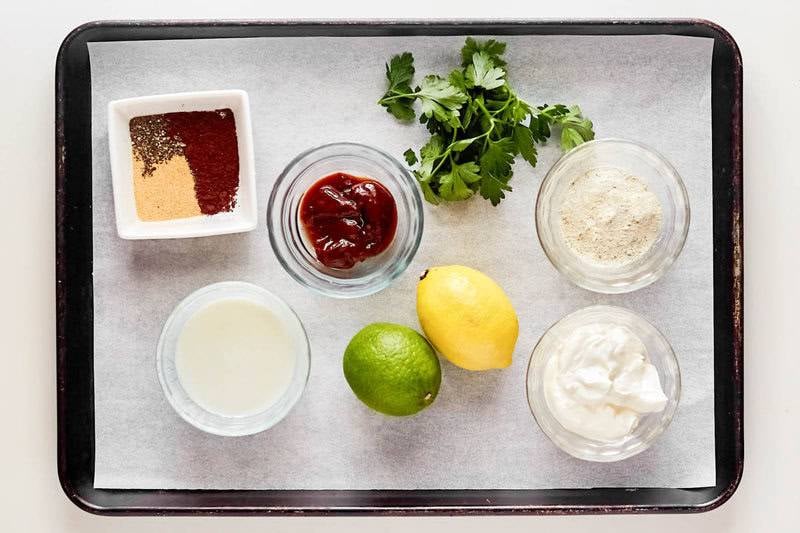 An overhead view of the ingredients for chipotle adobo ranch dip on a baking sheet. Pictured are bowls of sour cream, buttermilk, ranch mix, and chipotle peppers, along with a dish of spices, a fresh lemon, a lime, and a sprig of cilantro