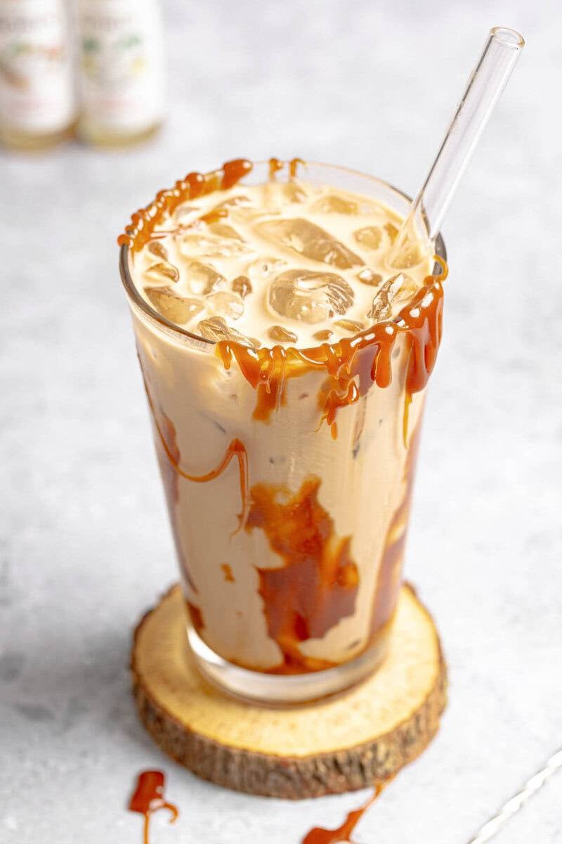 A finished Iced Golden Eagle latte in a tall glass with a heavy caramel drizzle inside and on top. Syrups in the background.