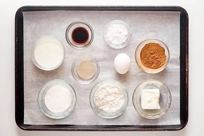 Ingredients for cinnamon rolls are measured into bowls on a tray: flour, sugar, butter, yeast, egg, cinnamon, and milk.