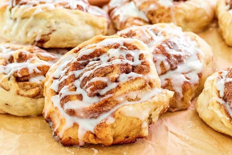 A close-up of a freshly baked cinnamon roll with icing, with several other rolls visible in the background on parchment paper.