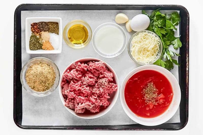 Ingredients for meatball parmigiana on a tray: ground beef, tomatoes, cheese, egg, breadcrumbs, milk, oil, and spices.