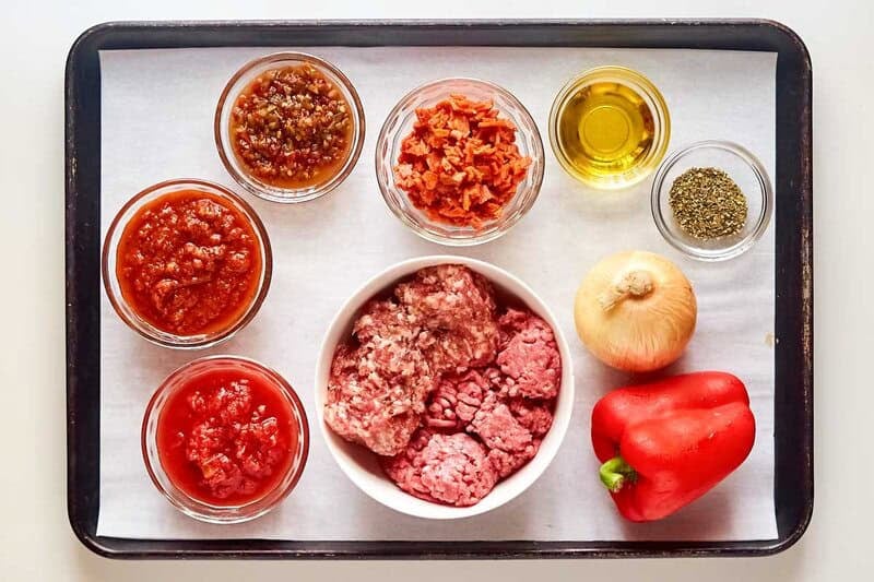 An overhead view of the ingredients for Olive Garden's spicy three-meat sauce on a baking sheet. Pictured are a bowl of raw ground beef and sausage, bowls of pepperoni, hot cherry peppers, marinara, crushed tomatoes, olive oil, and herbs, along with a whole onion and red bell pepper.