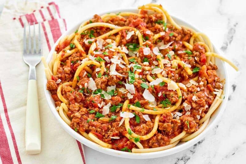 A white bowl filled with pasta tossed in a hearty, spicy three-meat sauce. The dish is garnished with shaved parmesan cheese and fresh parsley, with a fork and napkin resting nearby.