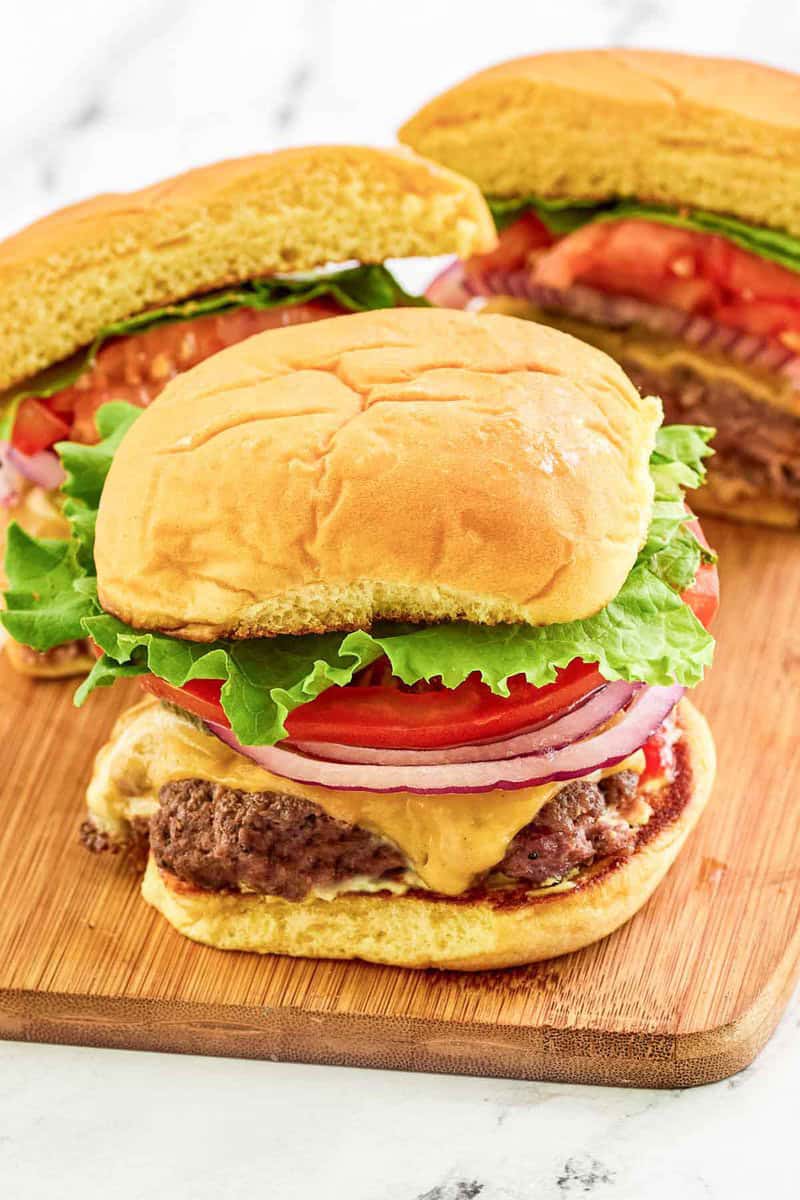 Two finished smash burgers with cheese, lettuce, tomato, and onion are arranged on a wooden board for serving.