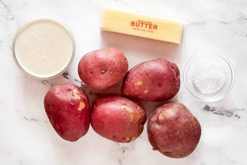 Overhead view of mashed potato ingredients: whole red potatoes, a stick of butter, salt, and heavy cream on a marble surface.