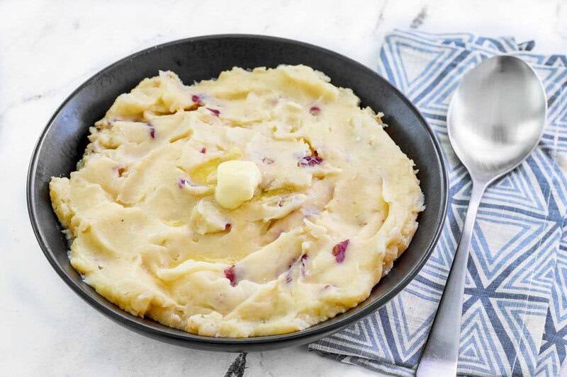 A black bowl of rustic mashed potatoes with red skin flecks and a pat of butter melting on top. A spoon and napkin are nearby.