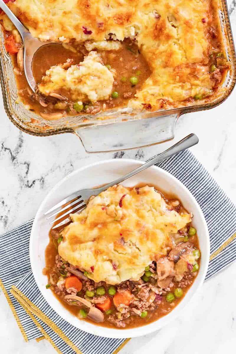 A white bowl filled with a serving of shepherd's pie. The pie has a saucy ground beef and vegetable filling, topped with golden mashed potatoes.