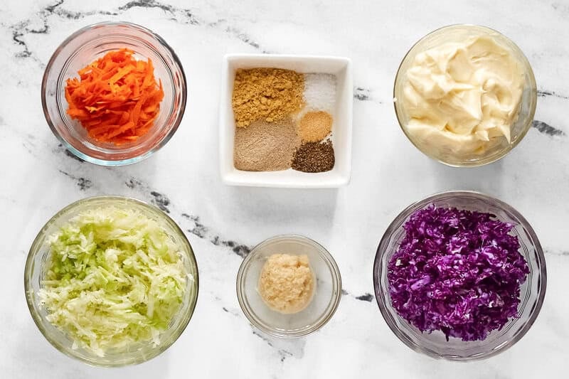 Overhead view of coleslaw ingredients: shredded cabbages, carrots, mayo, horseradish, and a dish of various dry spices.