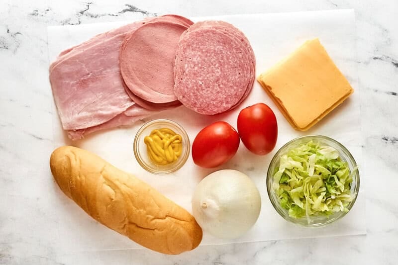 Ingredients for a Kmart sub sandwich laid out: sub roll, deli meats, American cheese, tomato, onion, lettuce, and mustard.