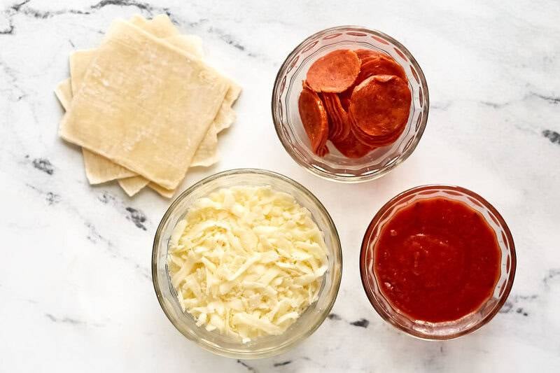 Overhead view of ingredients for pizza rolls: wonton wrappers, shredded mozzarella, pepperoni, and pizza sauce in bowls.