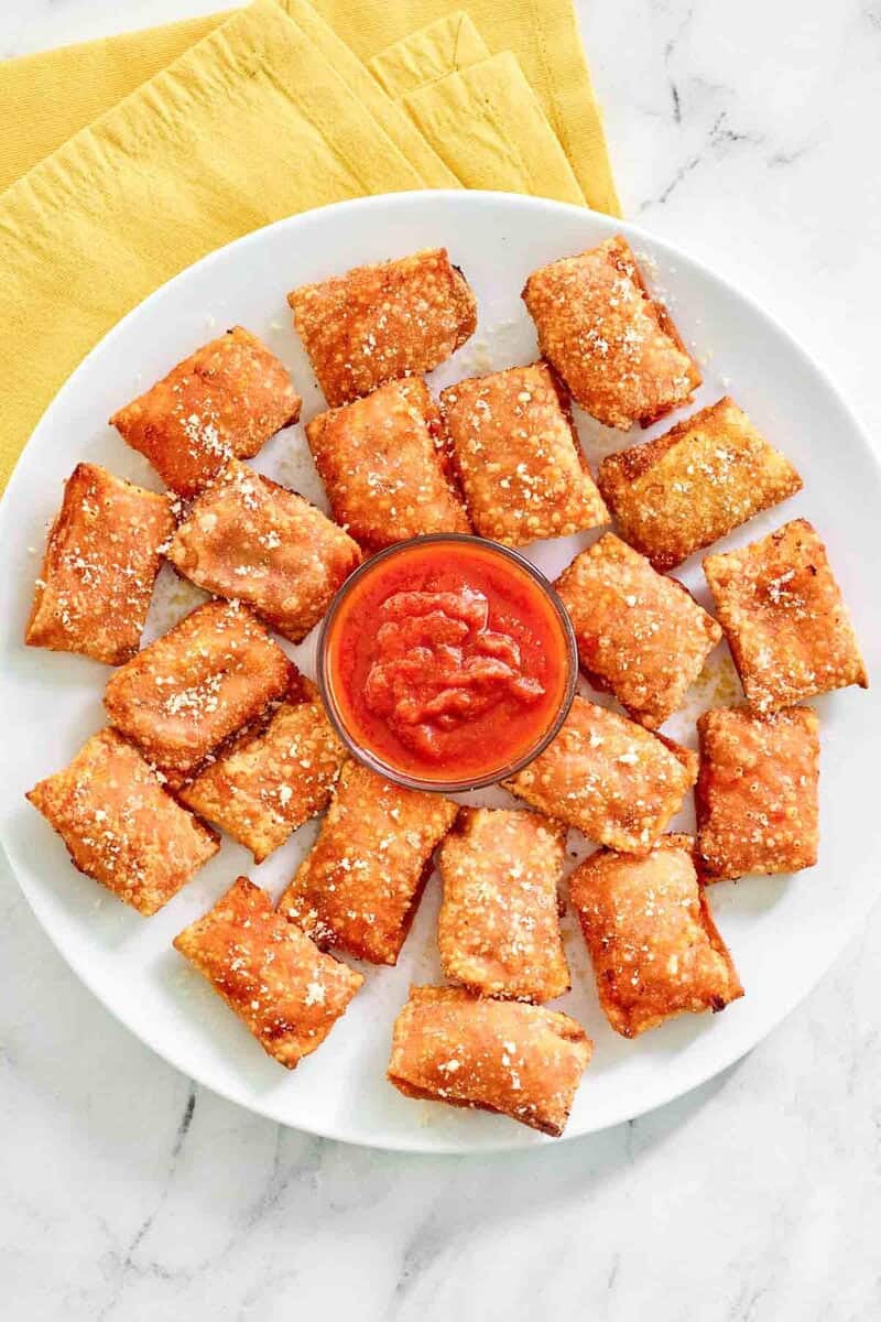 A plate of golden-fried pizza rolls, sprinkled with Parmesan, arranged around a small bowl of red dipping sauce.