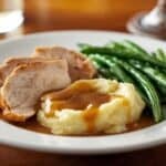 A restaurant-style plated turkey dinner with carved turkey slices, mashed potatoes with gravy drizzle, and green beans on a white plate with bright overhead lighting.