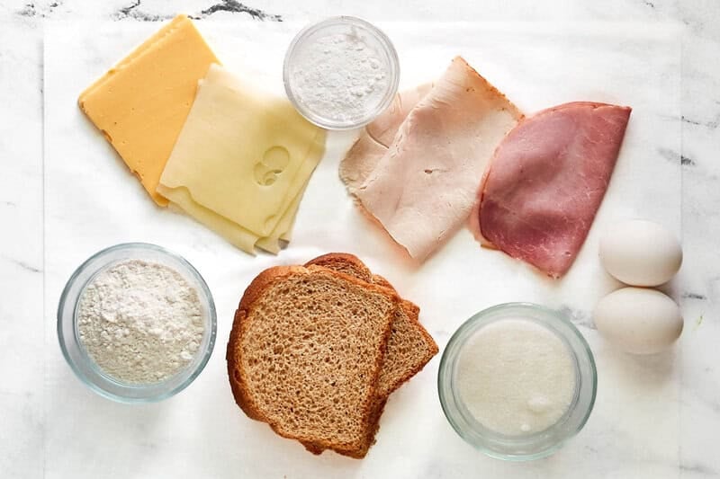 An overhead view of ingredients for a Monte Cristo: wheat bread, ham, turkey, two kinds of cheese, eggs, flour, and milk.