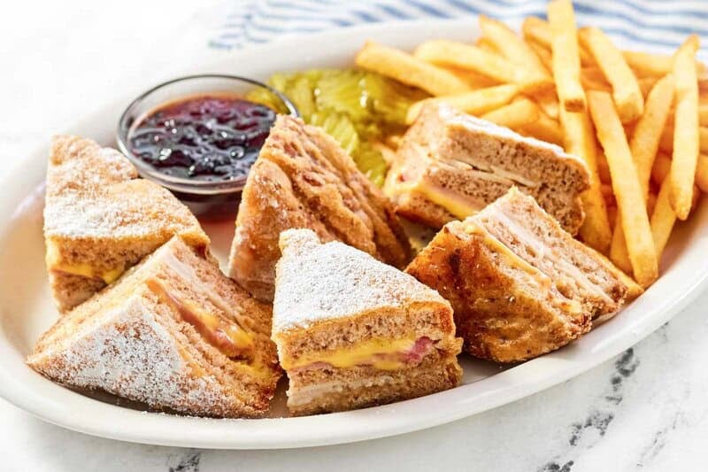 A platter with a Monte Cristo sandwich cut into triangles and dusted with sugar, served with fries, pickles, and jam.