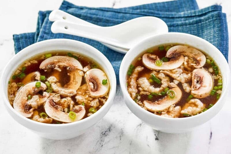 Two white bowls of Japanese clear soup, garnished with sliced mushrooms and scallions, sit next to white soup spoons.