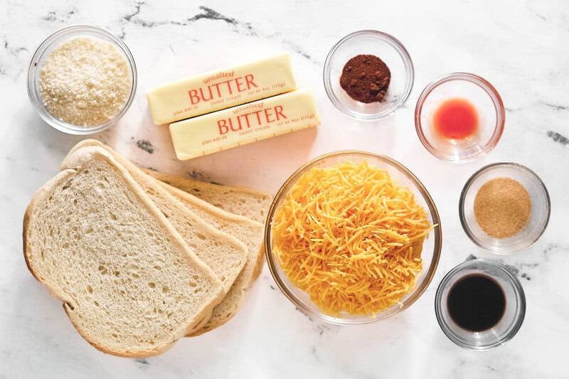 Overhead view of ingredients: bread slices, sticks of butter, shredded cheddar, grated Romano, and spices in bowls.