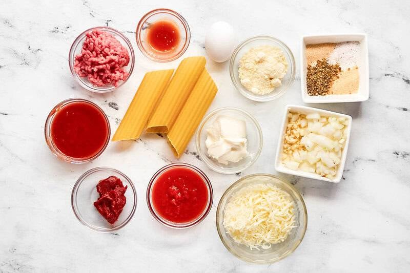 Overhead view of manicotti ingredients: pasta shells, ground beef, tomato sauce, ricotta, mozzarella, parmesan, egg, and spices.
