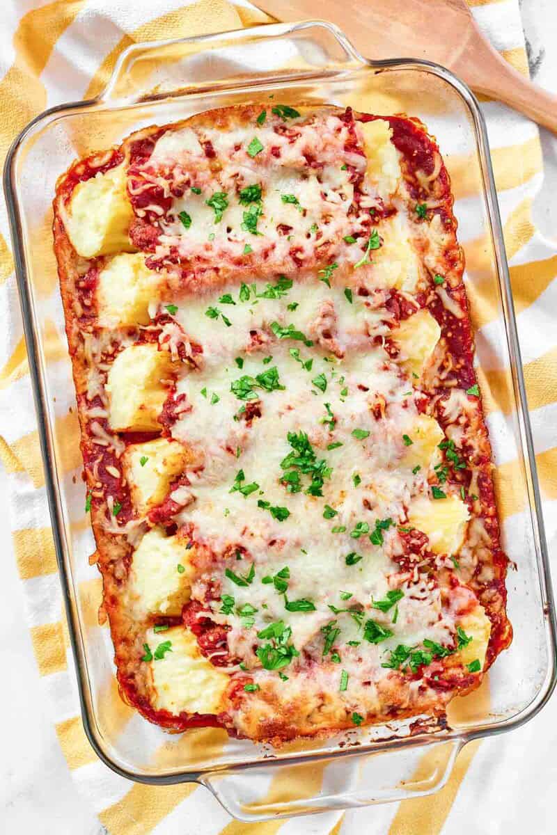 A baked casserole of four cheese manicotti in rich meat sauce, topped with melted cheese and parsley, on a yellow and white towel.