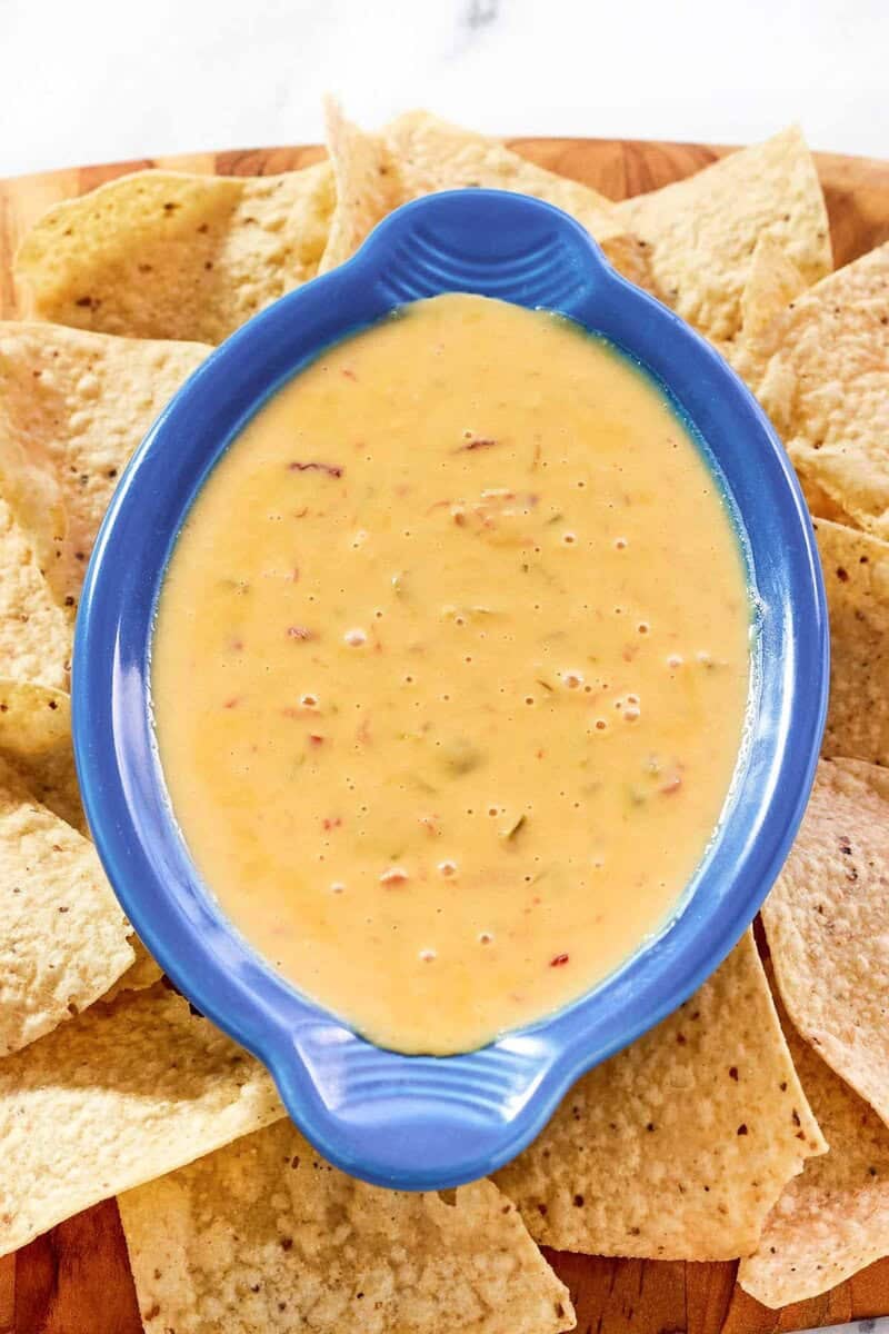 Top-down view of spicy queso dip in a blue bowl, surrounded by corn tortilla chips on a wooden serving platter.