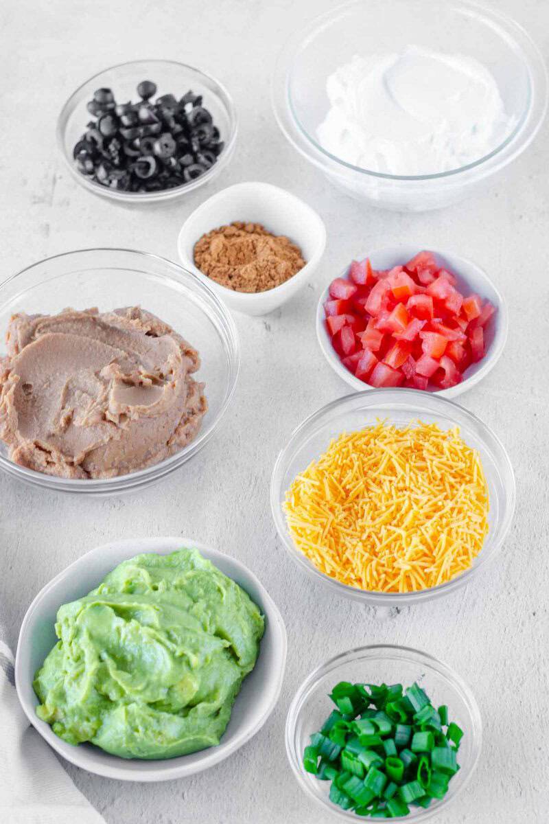 Ingredients for 7-layer dip arranged in bowls: bean dip, guacamole, sour cream, cheese, tomatoes, onions, olives, and spices.ace.