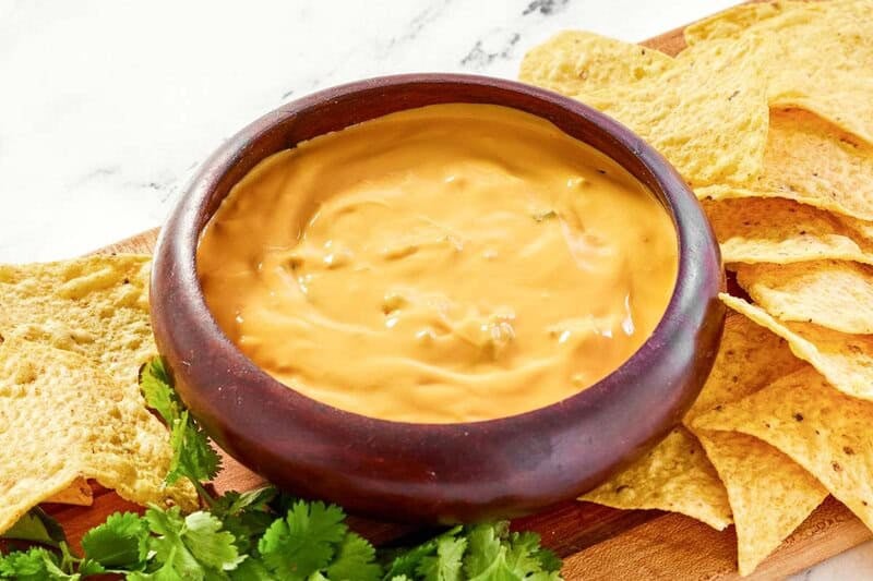 Close up of smooth Green Chile Queso in a bowl served with crispy tortilla chips on a wooden board.