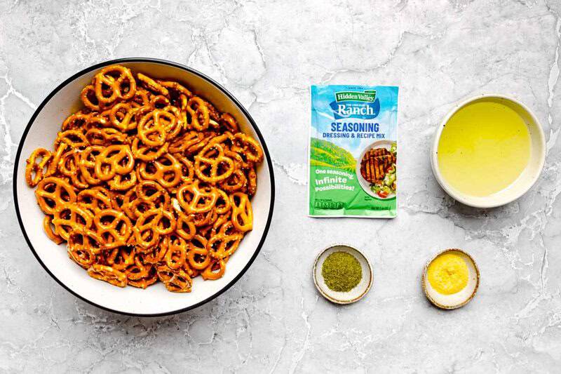 Overhead view of ingredients: a large bowl of pretzels, a packet of ranch seasoning, oil, dill weed, and garlic powder.