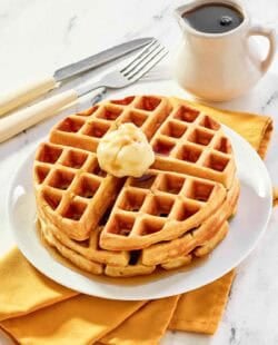 Three homemade waffles stacked on a plate and topped with butter and syrup.