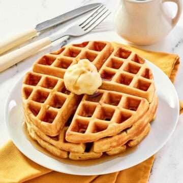 Three homemade waffles stacked on a plate and topped with butter and syrup.