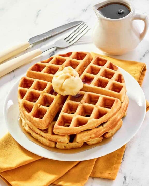 Three homemade waffles stacked on a plate and topped with butter and syrup.
