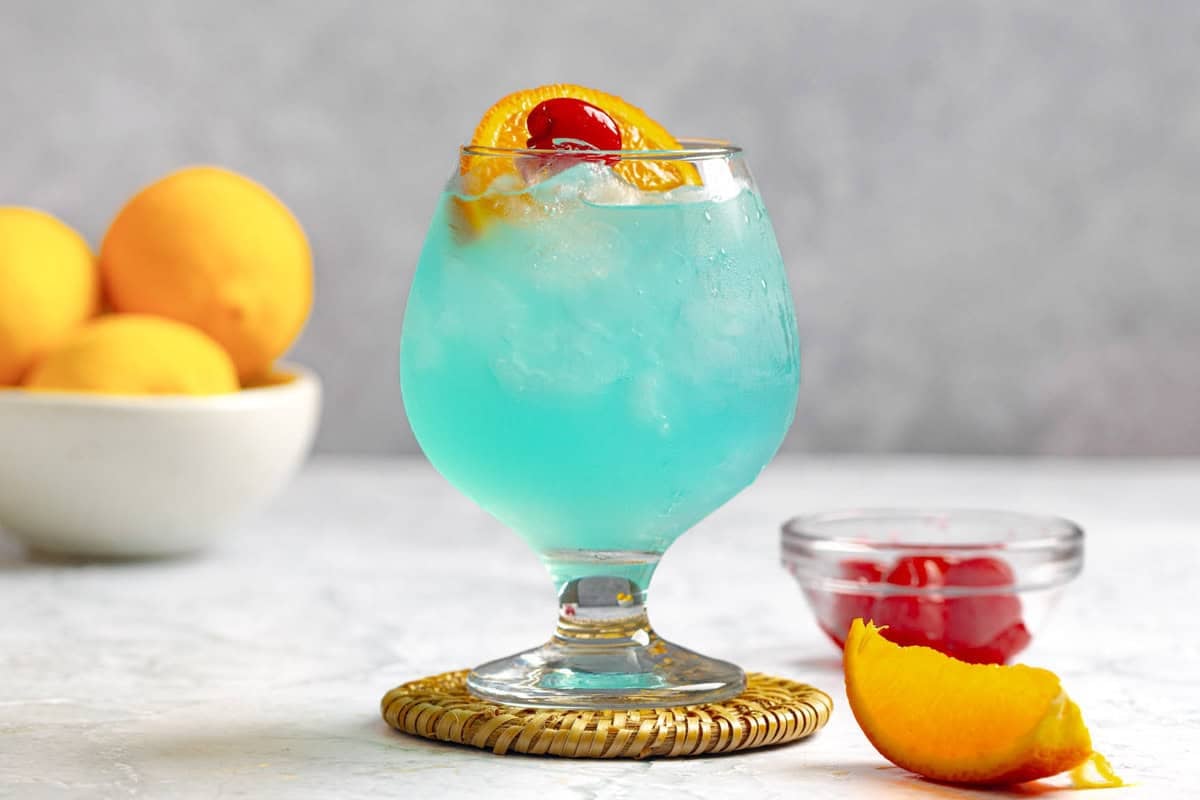 A rum, blue curacao, and lemonade cocktail, lemons in a bowl, and a bowl of cherries.