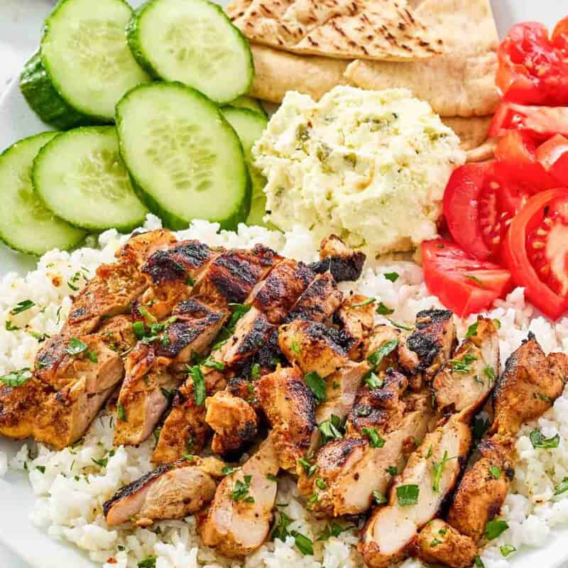 A plate of harissa chicken, rice, feta spread, cucumber, tomato, and pita bread.