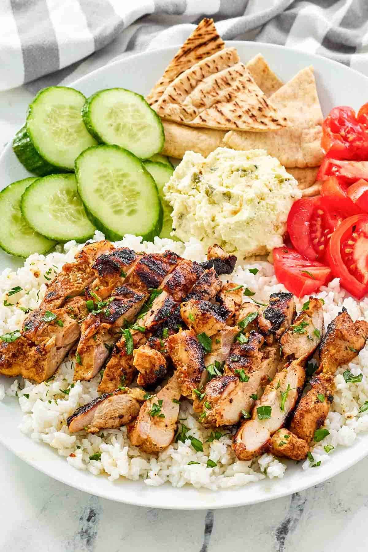 A plate of harissa chicken, rice, feta spread, cucumber, tomato, and pita bread.