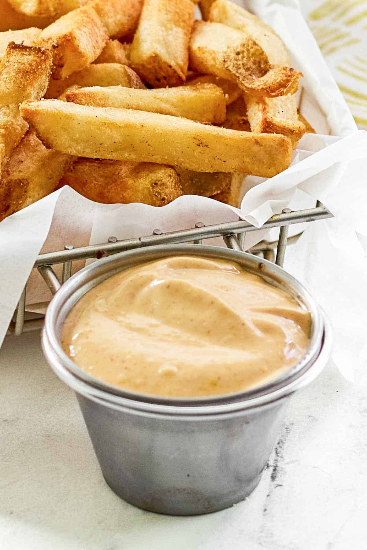 French fries in a parchment lined wire basket and a small cup of creamy seasoned dipping sauce.
