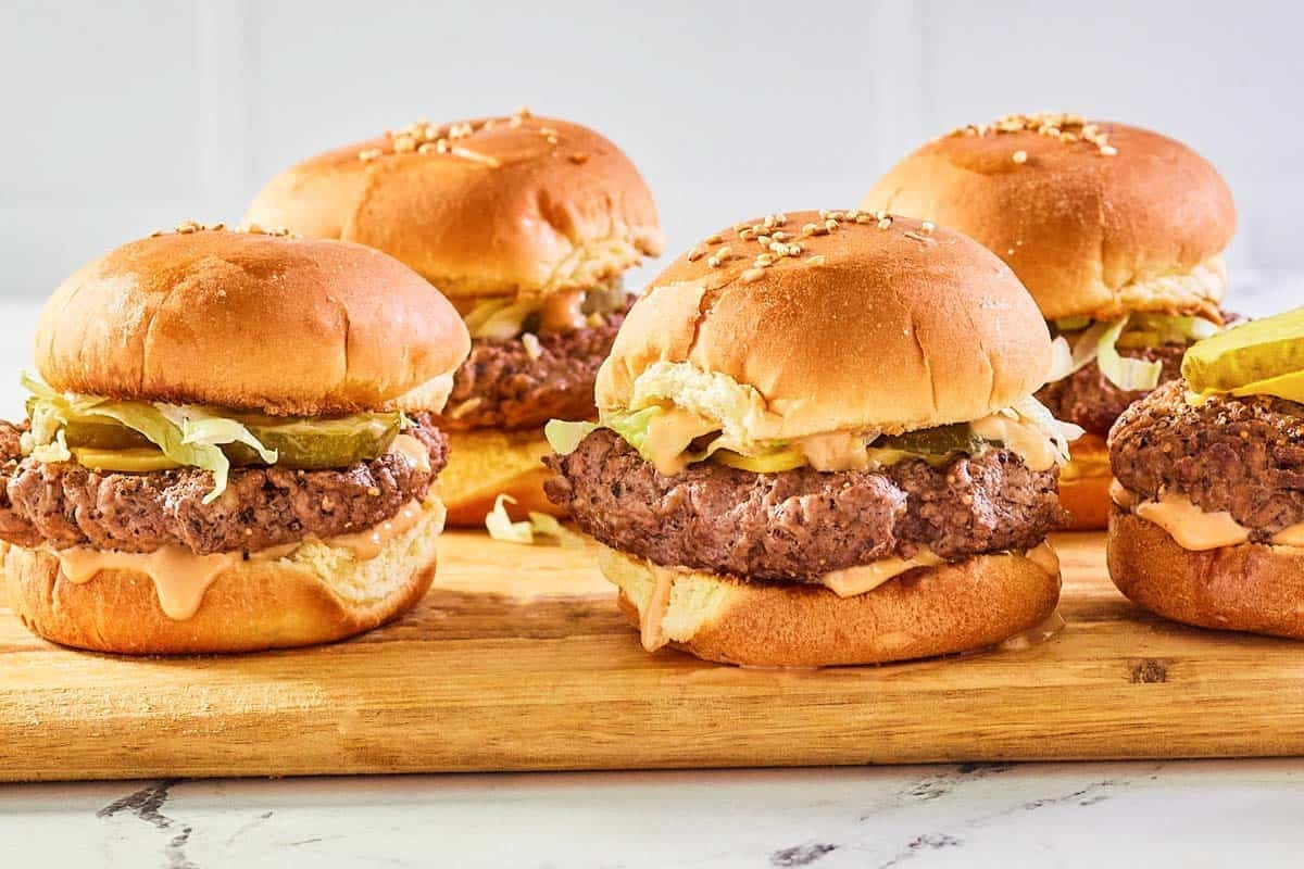 Ground beef sliders with sauce, cheese, pickles, onions, and lettuce on sesame seed buns.